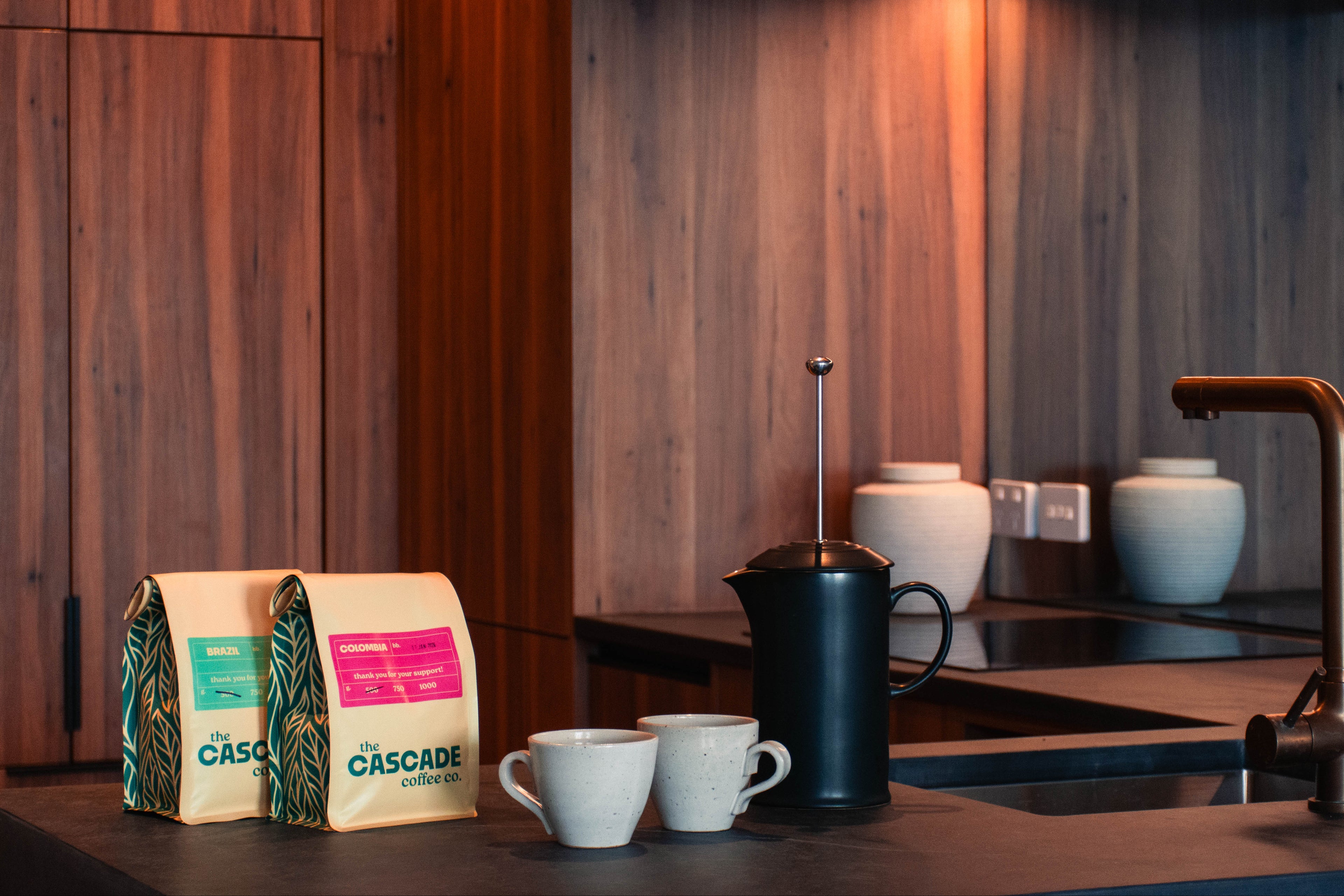 Two Cascade coffee bags on a kitchen counter with a coffee maker and cups.
