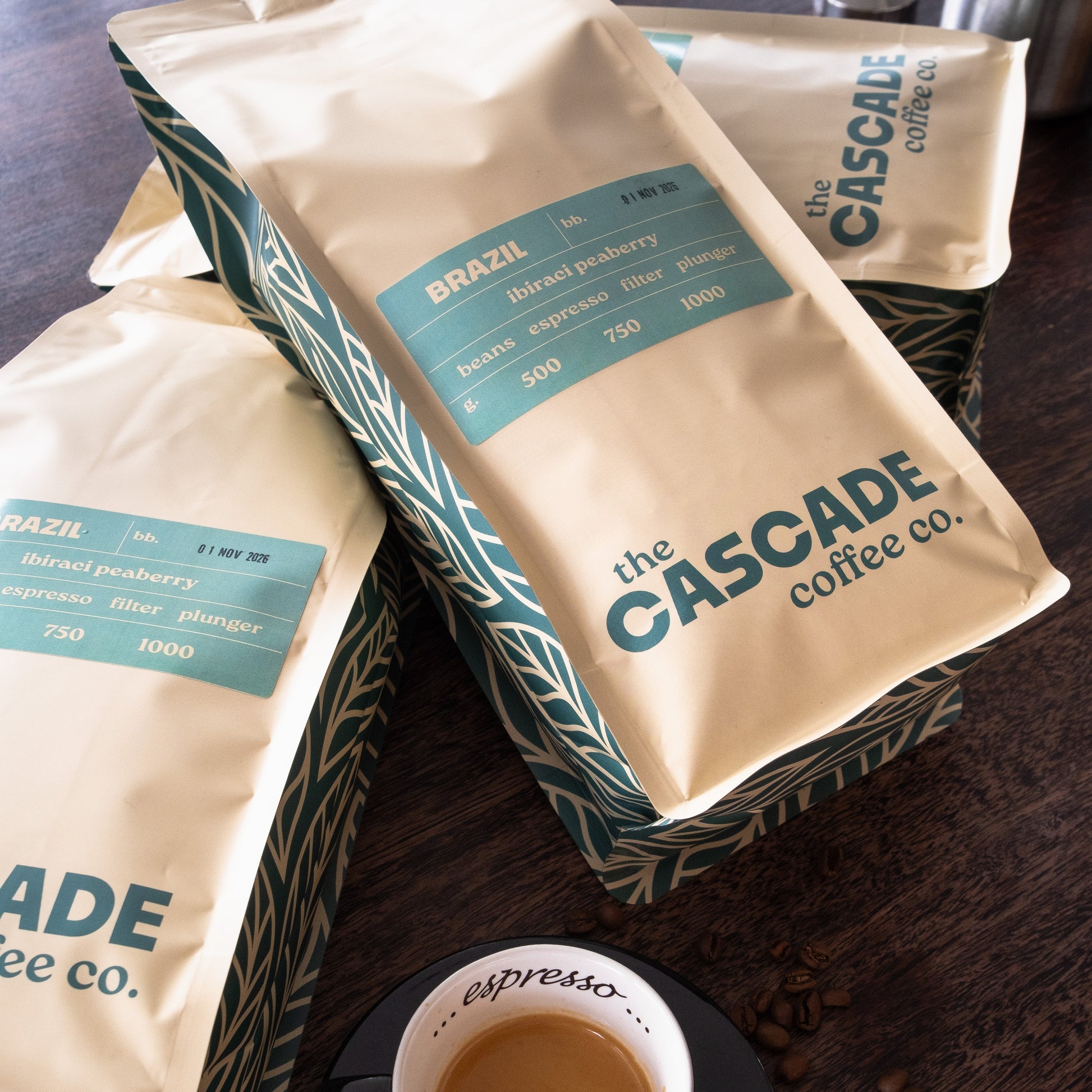 Coffee bags from The Cascade Coffee Co. with a cup of coffee on a wooden surface.